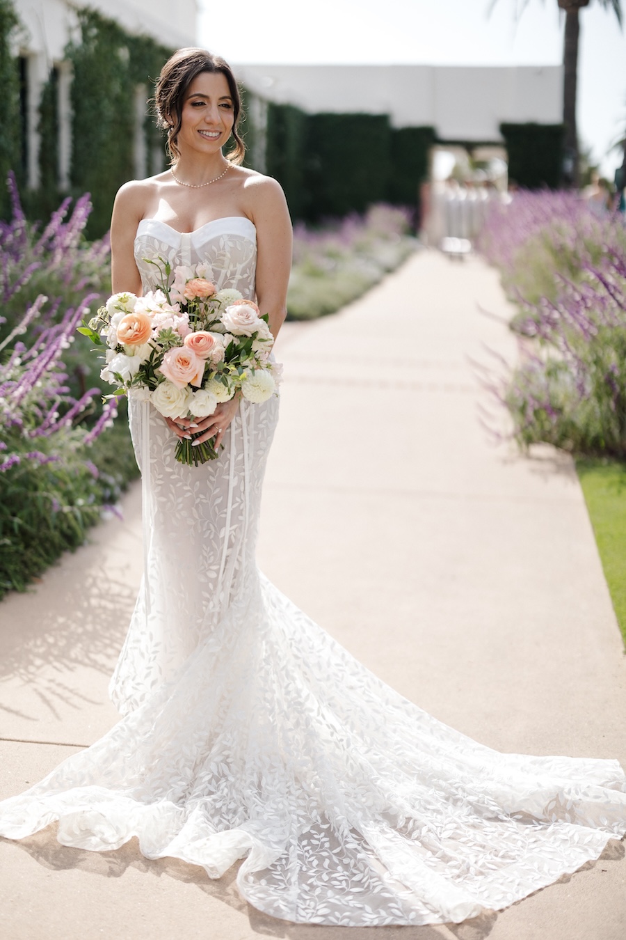 Bride holding bouquet standing in path lined with lavender bushes. 